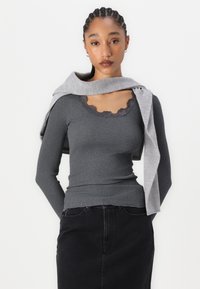 Haut à manches longues côtelé gris avec une dentelle au niveau de l'encolure, associé à un pull gris clair drapé sur les épaules. Jupe noire.