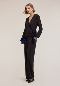 Top avvolgente nero a maniche lunghe con dettaglio a fiocco, abbinato a pantaloni a gamba larga. Tessuto liscio, scollatura a V profonda e décolleté neri eleganti.