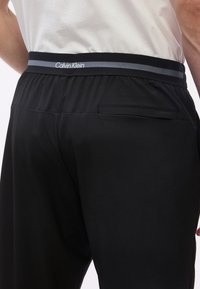 Άνδρας που φοράει μαύρο παντελόνι με ζώνη μέσης με την επωνυμία Calvin Klein και τσέπη στη δεξιά πίσω πλευρά, σε συνδυασμό με άσπρο πουκάμισο.