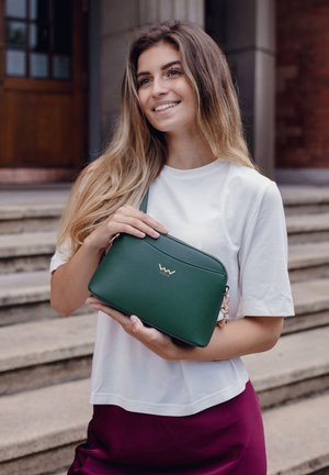 Zelená texturovaná kožená crossbody taška se zlatým logem, zipovým uzávěrem a odnímatelným popruhem, kterou drží osoba v bílé košili a bordó sukni.