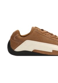 Zapatillas de ante marrón con acentos en cuero blanco, puntera redonda, superficie texturizada, suela de goma plana y detalles cosidos a lo largo de los lados.