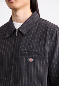 Černý sako s proužky a zipovým zapínáním, s límečkem košile a náprsní kapsou se značkou Dickies. Bavlněná tkanina, strukturovaný povrch.