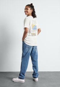 Ljusgrå t-shirt med en färgglad grafik av en sneaker på baksidan, matchad med lösa blå denimjeans och vita sneakers.