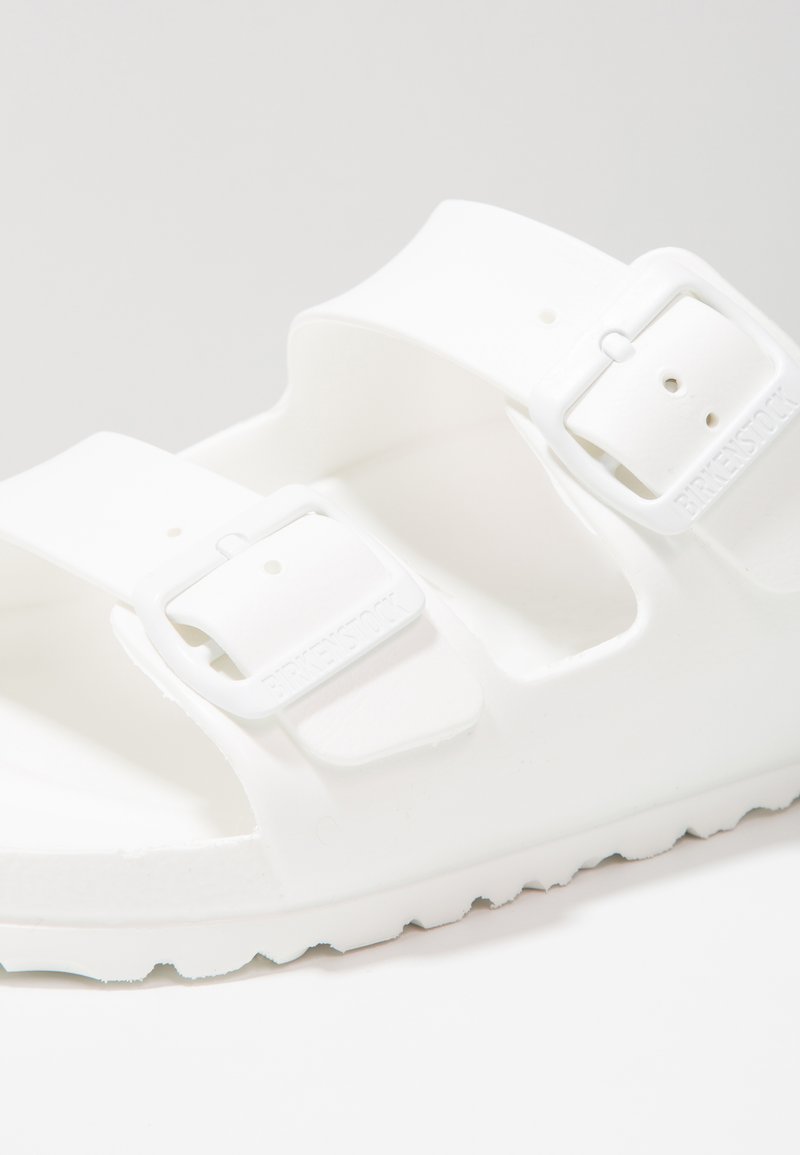 Infradito bianche con due cinturini regolabili, texture liscia e suola resistente. Presenta un logo in rilievo sul cinturino, progettato per comfort e supporto.