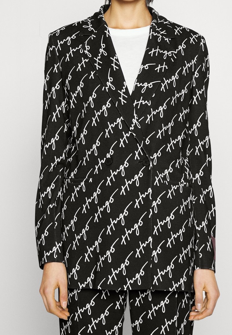 Blazer noir croisé avec "Hugo" en écriture cursive blanche imprimé en diagonale, porté sur un top blanc, associé à un pantalon imprimé assorti.