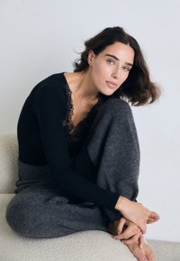 Kvinna med ljus hy och blå ögon sitter i lotusställning på en vit soffa, iförd en svart långärmad topp med spets och grå byxor.