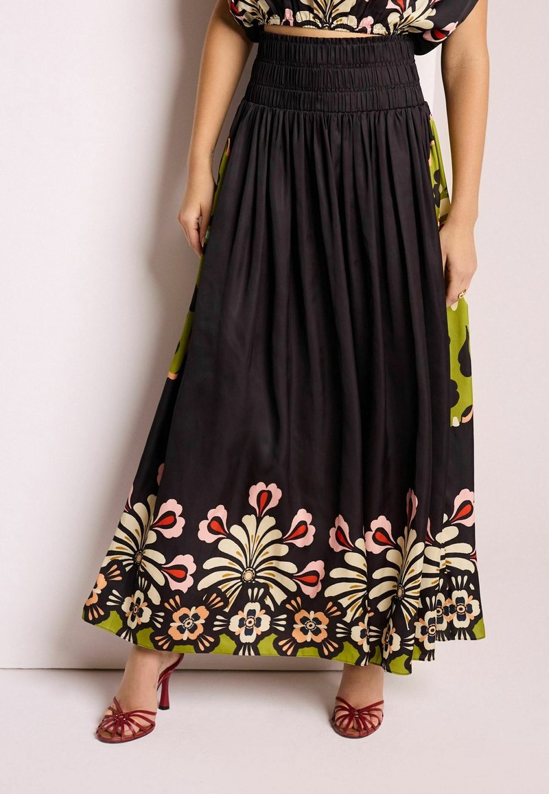 Femme portant une longue jupe noire à grands motifs floraux colorés et des sandales à talons hauts rouges à brides, se tenant contre un mur blanc.