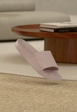 Miękki różowy, otwarty na palce klapek leżący obok drewnianego, okrągłego stolika z poukładanymi książkami w minimalistycznym salonie.