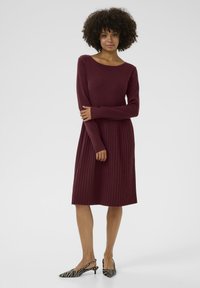 Burgundská pletená šaty s priliehajúcim korzetom a skladanou sukňou. Dlhé rukávy. V kombinácii s čierno-bielymi pruhovanými slingback topánkami.