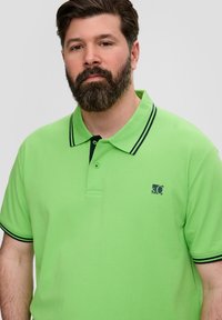 Zielona koszulka polo z fakturowanej tkaniny, z czarnym kontrastowym kołnierzykiem i mankietami oraz małym logo na lewej piersi.