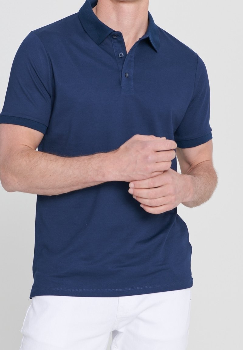 Homme portant un polo à manches courtes bleu marine et un pantalon blanc, mains jointes à la taille devant un fond uni.