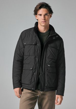 Veste noire isolée avec un motif en losanges, plusieurs poches avant, col montant et fermeture éclair. Portée sur un pull en maille vert foncé.