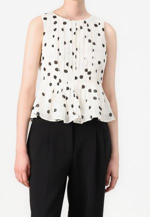 Woman wearing sleeveless white blouse with black polka dots and pleats, paired with high-waisted black trousers against a plain background.