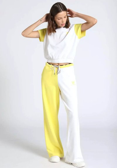 Donna che sistema i capelli, indossando una maglietta a maniche corte bianca e gialla a blocchi di colore e pantaloni a gamba larga divisi nei colori abbinati, con scarpe bianche.