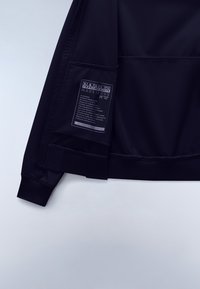 Rękaw czarnej kurtki i obszar biodrowy z prostokątną białą metką zawierającą nazwę marki "Napapijri" oraz tekst o tkaninie i zrównoważonym rozwoju.