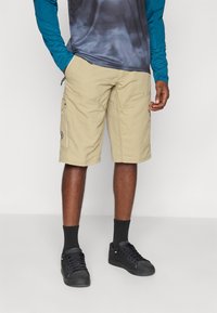 Shorts cargo beiges avec poches latérales, fabriqués en tissu léger, associés à des baskets noires et des chaussettes sombres, avec un design uni.