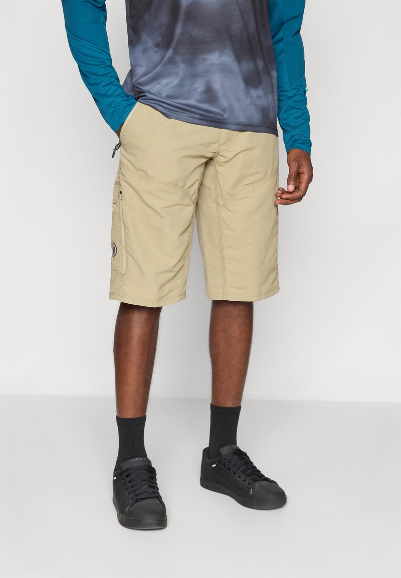 Shorts cargo beiges avec poches latérales, fabriqués en tissu léger, associés à des baskets noires et des chaussettes sombres, avec un design uni.