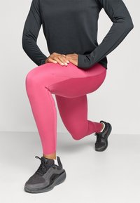 Tilpassede lyserøde leggings med struktureret sideløsning og en glat finish, parret med en langærmet sort top og sorte atletiske sko.