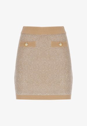 Mini-jupe beige en maille avec ceinture côtelée, deux poches avant ornées de boutons dorés et motif texturé sur le tissu.