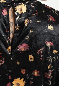 Gros plan sur un tissu noir avec un motif floral jaune, rouge et rose, présentant un col en trou de serrure boutonné dévoilant une partie d'une poitrine tatouée.