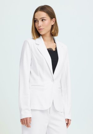 Õlgadeni juukseline naine, kes kannab valget bleiserit ja pükse musta pitsäärega topi kohal, vaadates kergelt küljele.