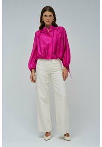 Donna che indossa una blusa di seta fucsia con maniche lunghe e gonfie, pantaloni bianchi a vita alta e a gamba larga, e scarpe bianche con punta, in piedi contro uno sfondo grigio.