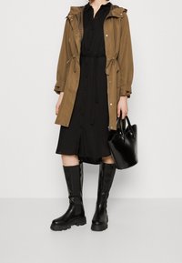 Giacca impermeabile marrone con coulisse sopra un vestito nero con bottoni. Stivali di gomma neri al ginocchio e una borsa nera lucida completano il look.