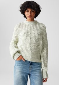 Maglione lavorato a maglia verde chiaro con collo alto, texture a coste e maniche ampie. Indossato con jeans blu, parzialmente infilato in vita.