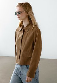 Giacca in suede marrone con colletto, bottoni frontali e taschino sul petto, abbinata a jeans azzurri a vita alta e occhiali da sole rotondi.