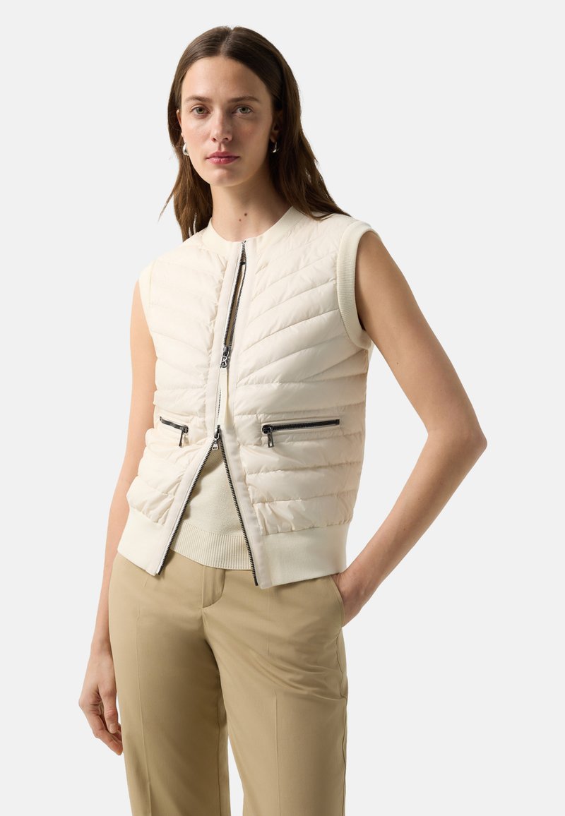 Vrouw draagt mouwloos gewatteerd ivoorkleurig vest met voorrits en zakken, beige broek, staat met één hand in de zak tegen een effen achtergrond.