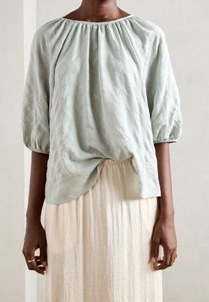 Blouse texturée vert clair avec un large col et des détails froncés ; associé à une jupe fluide crème. Manches trois quarts.