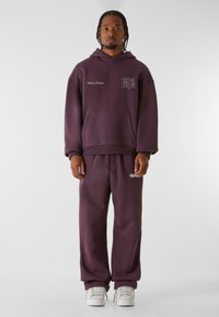 Dunkel lila Kapuzenpullover mit Fronttasche und Logo auf der Brust, kombiniert mit passenden Jogginghosen. Glatte Textur, lockere Passform und elastische Bündchen.