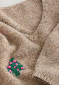 Beige uldtrøje med V-udskæring og en blød tekstur, der har et grønt broderet blomstermotiv med lyserøde accenter på fronten.