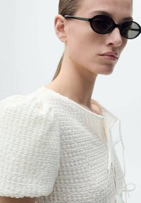 Top bianco con texture, maniche corte a sbuffo, tessuto leggero e apertura frontale legata con sottili corde bianche. Occhiali da sole ovali neri.