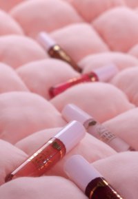 Vijf lipgloss-tubes met witte doppen en gouden accenten liggen op een zachte, lichtroze, gewatteerde stof. De kleuren zijn transparant, roze en bruin.