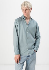 Camisa azul clara de tejido suave, con cuello tipo solapa, botón en el frente y mangas largas. Lleva puesta unos pantalones de mezclilla claros.
