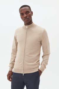 Bežs kreklu ar rāvējslēdzēju un augstu apkakli, pieguļošs dizains, ribotas piedurknes un apakšmala. Mīksta tekstūra, piemērota kārtu veidošanai.