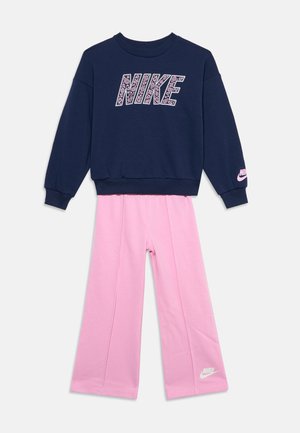 Granatowa bluza Nike z różowym logo paisley w zestawieniu z jasnoróżowymi szerokimi spodniami Nike z białym logo przy krawędzi.