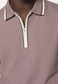 Pohodlná polokošile s krátkým rukávem z mauvové bavlny. Má límec na zip se zelenými a bílými pruhy. Hladká textura a minimalistický design.