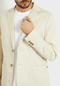 Blazer de algodón beige con solapas estructuradas, dos botones y un bolsillo en el pecho. Presenta puños abotonados y una textura suave y ligera.