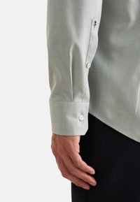 Camicia verde chiaro con un tessuto texture. Presenta un polsino con bottoni e un piccolo dettaglio ricamato a forma di rosa sulla manica.