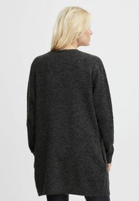 Femeie blondă îmbrăcată într-un pulover supradimensionat de tricot gri închis, vazută din spate pe un fundal simplu, luminos.
