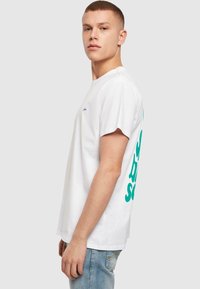 Hvid bomulds t-shirt med rund hals, der har et turkist grafisk design på ryggen. Korte ærmer og afslappet pasform. Lyseblå jeans.