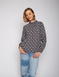 Schwarz-weiße Bluse mit gemusterten runden Designs, hochgeschlossenem Hals und gerafften Ärmelbündchen, kombiniert mit hellblauen, verwaschenen Jeans.