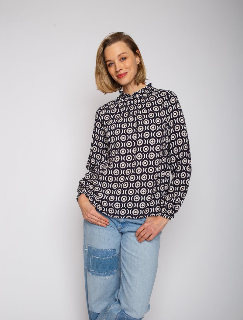 Schwarz-weiße Bluse mit gemusterten runden Designs, hochgeschlossenem Hals und gerafften Ärmelbündchen, kombiniert mit hellblauen, verwaschenen Jeans.