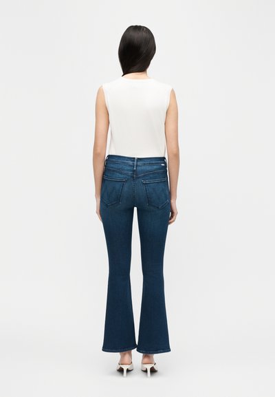 Γυναίκα που φοράει λευκό μπλουζάκι χωρίς μανίκια, ψηλόμεσα σκούρα μπλε τζιν καμπάνες και λευκά σανδάλια με τακούνι, γυρισμένη με την πλάτη σε απλό φόντο.