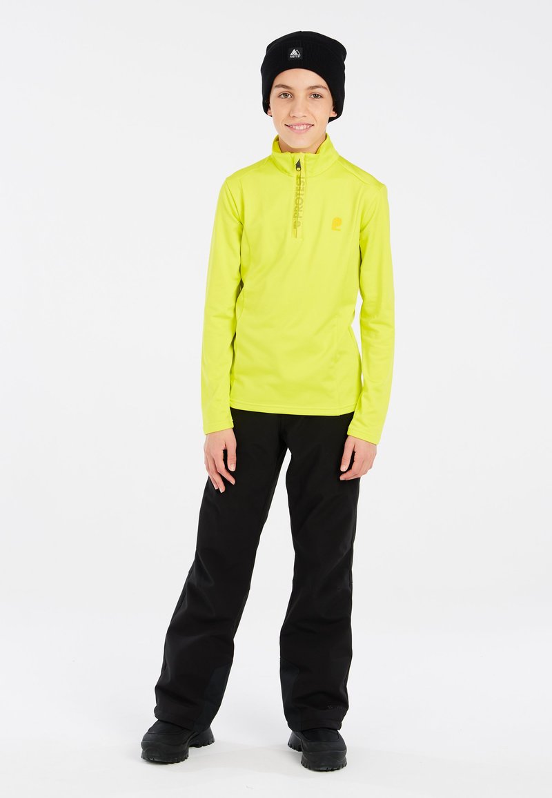 Maglia termica a maniche lunghe di un brillante giallo con zip e logo, accompagnata da pantaloni neri larghi e stivali neri robusti. Texture liscia.