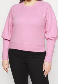 Różowa bluza z raglanowymi rękawami, z okrągłym dekoltem i skróconym hemem. Materiał wydaje się miękki, o gładkiej fakturze i minimalnych detalach szwów.