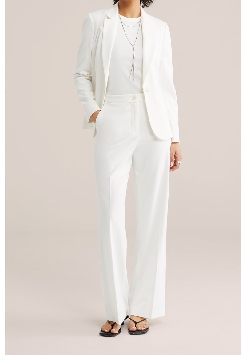 Femme portant un costume blanc avec blazer et pantalon à jambes larges, associé à des sandales noires à bout ouvert et un long collier en argent.