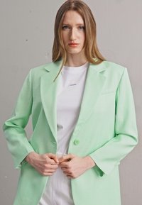 Femme avec de longs cheveux lisses portant un blazer vert clair sur un t-shirt blanc, se tenant devant un fond gris uni.